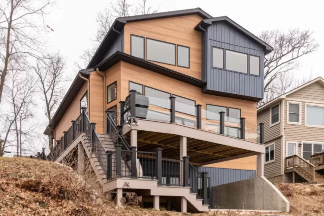 Modern Lakefront Home with Deck and Stairs | Pennings & Sons Modern lakefront home exterior with multi-level deck, glass railing, exterior stairs, black railings, wood-look siding, and dark vertical siding