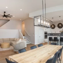 Open concept interior with dining table, living room seating, staircase, kitchen island, and modern pendant lights