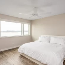 Primary bedroom with white bed, white ceiling fan, wood-look flooring, neutral walls, and wide lake-facing windows