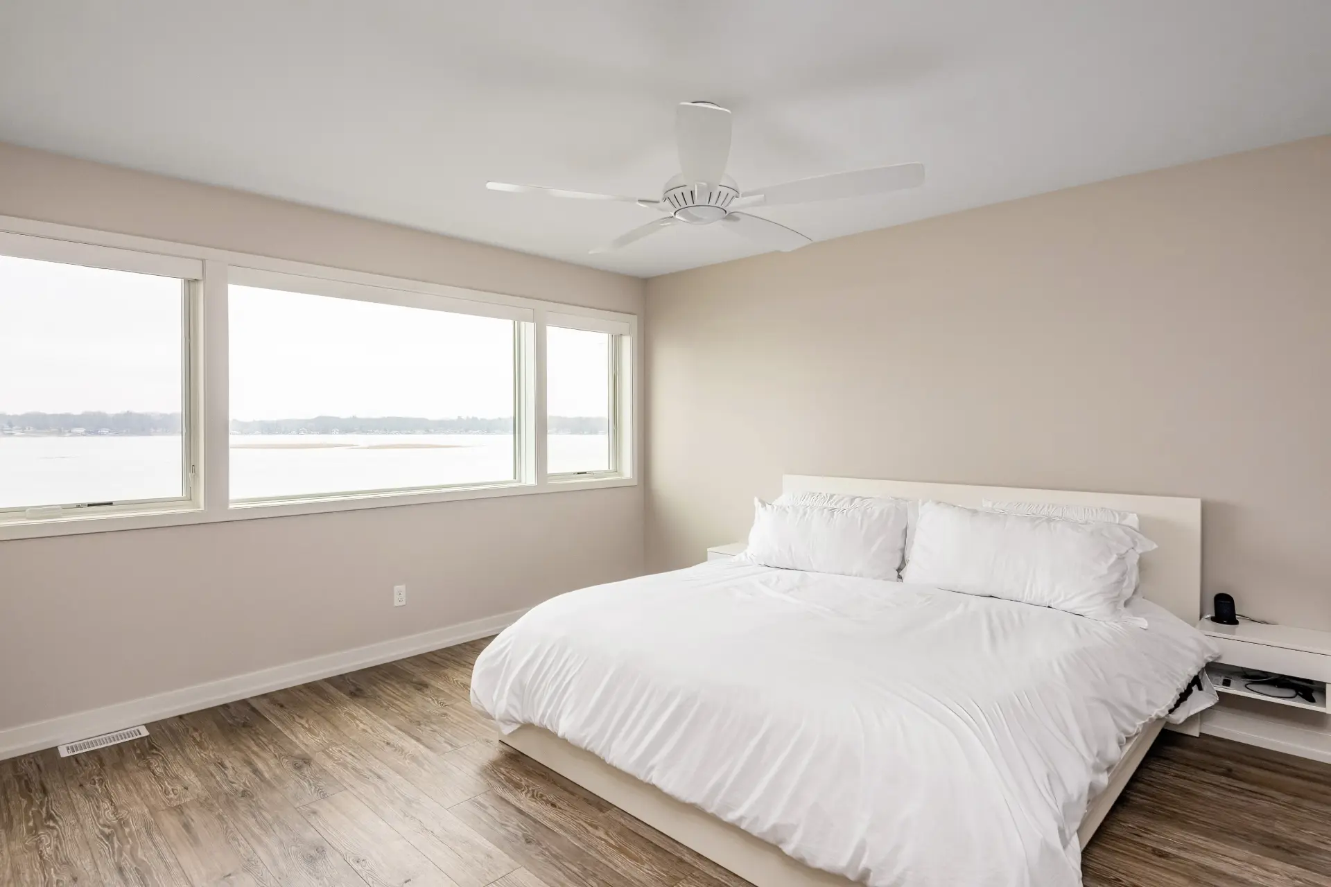 Primary bedroom with white bed, white ceiling fan, wood-look flooring, neutral walls, and wide lake-facing windows