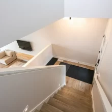View down the staircase toward the entry and living room with wood stair treads, white half wall, black rug, and wall-mounted TV