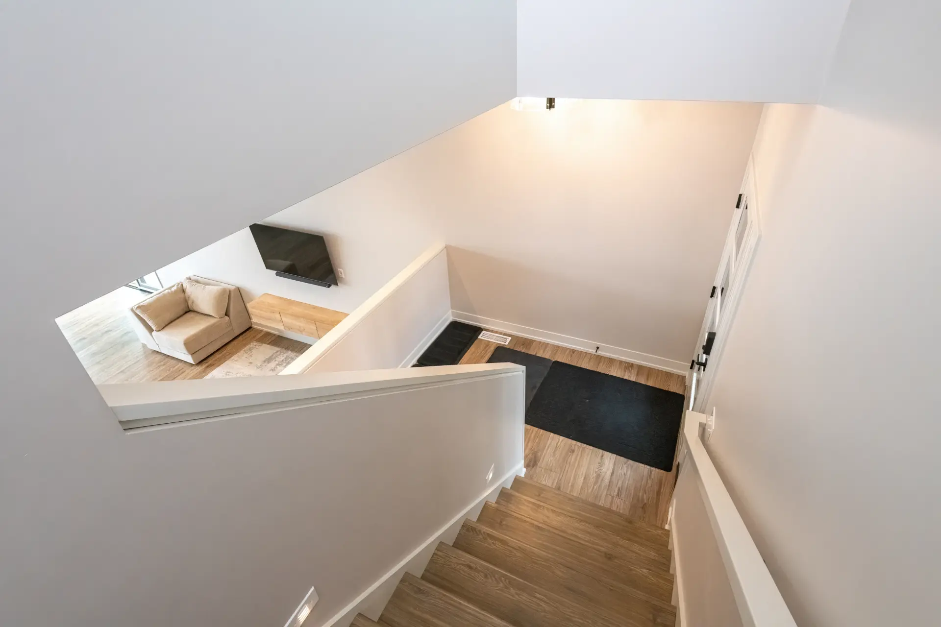 View down the staircase toward the entry and living room with wood stair treads, white half wall, black rug, and wall-mounted TV