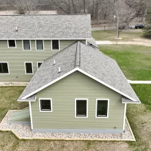 Aerial rear exterior view of green home with gray shingle roof, white trim windows, walkways, and surrounding yard