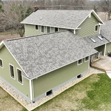 Aerial side view of green home with gray shingle roof, small deck, skylights, white trim, and landscaped perimeter