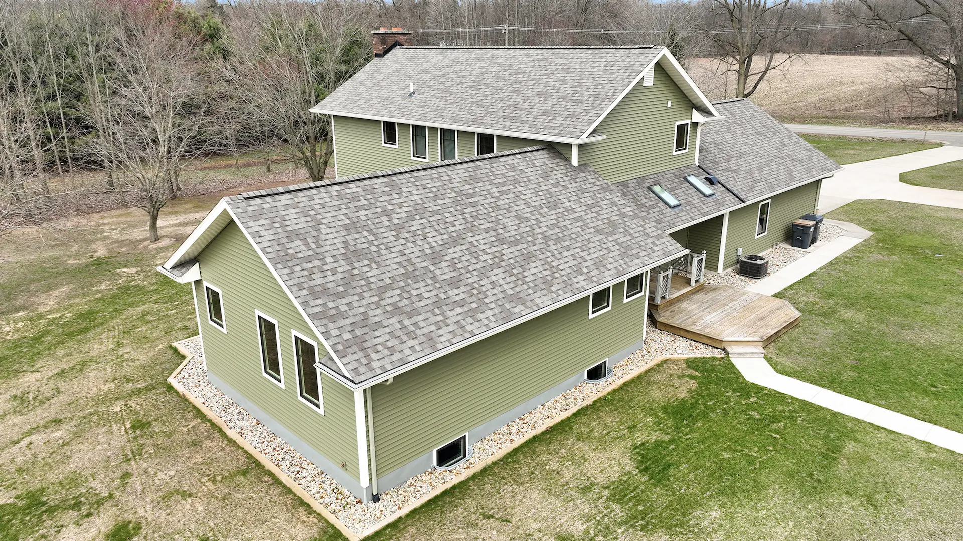 Aerial side view of green home with gray shingle roof, small deck, skylights, white trim, and landscaped perimeter