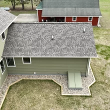 Aerial view of green home exterior with gray shingle roof, white trim windows, rock landscaping, and open lawn