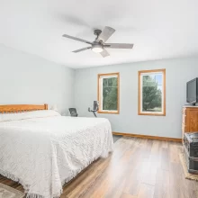 Bedroom with white bedding, light blue walls, wood flooring, ceiling fan, wood trim windows, dresser, and TV