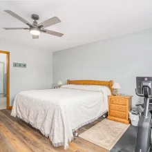 Bedroom with white bedding, wood furniture, exercise equipment, ceiling fan, wood flooring, and ensuite bathroom access