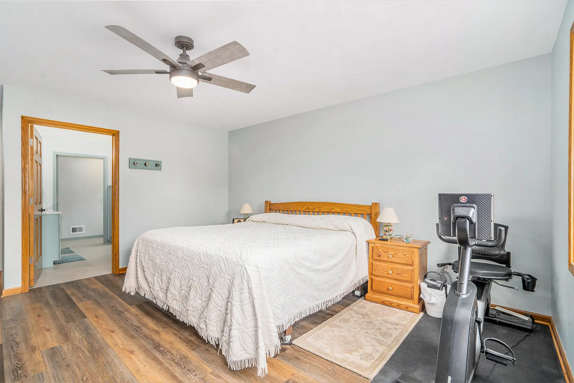 Bedroom with white bedding, wood furniture, exercise equipment, ceiling fan, wood flooring, and ensuite bathroom access
