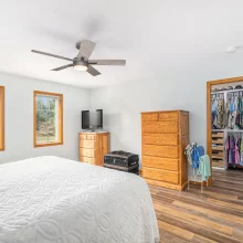 Bedroom with white bedding, wood furniture, ceiling fan, two windows, wood flooring, TV, and closet access