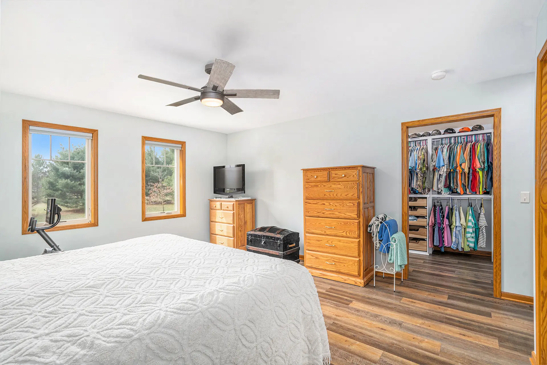 Bedroom with white bedding, wood furniture, ceiling fan, two windows, wood flooring, TV, and closet access