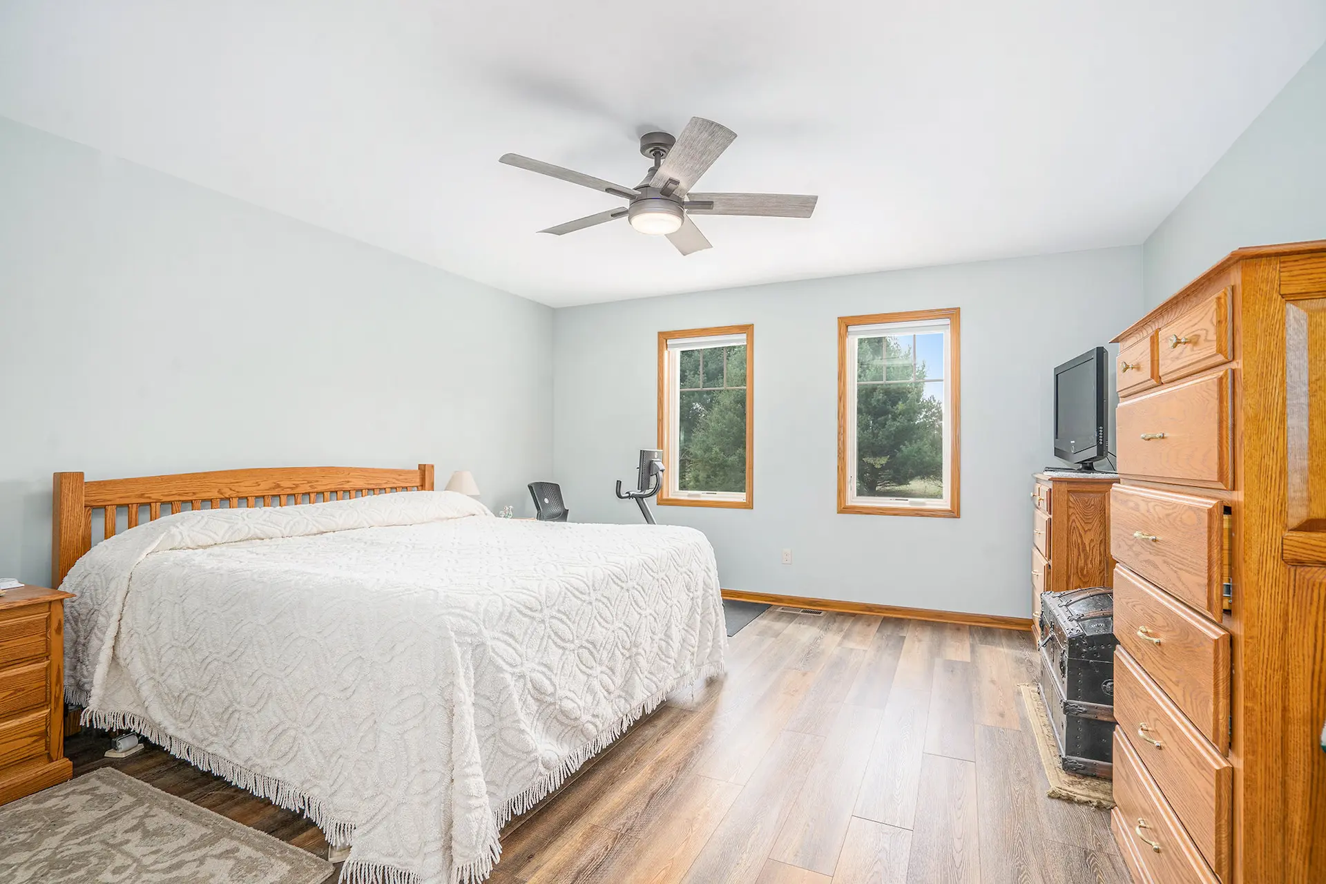 Bedroom with white bedding, light blue walls, wood flooring, ceiling fan, wood trim windows, dresser, and TV