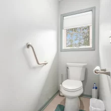 Compact half bathroom with toilet, window, grab bars, towel bar, light walls, and blue-green trim details
