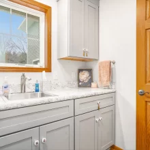 Laundry room utility sink with gray shaker cabinets, stone-look countertop, wood window trim, and wood door