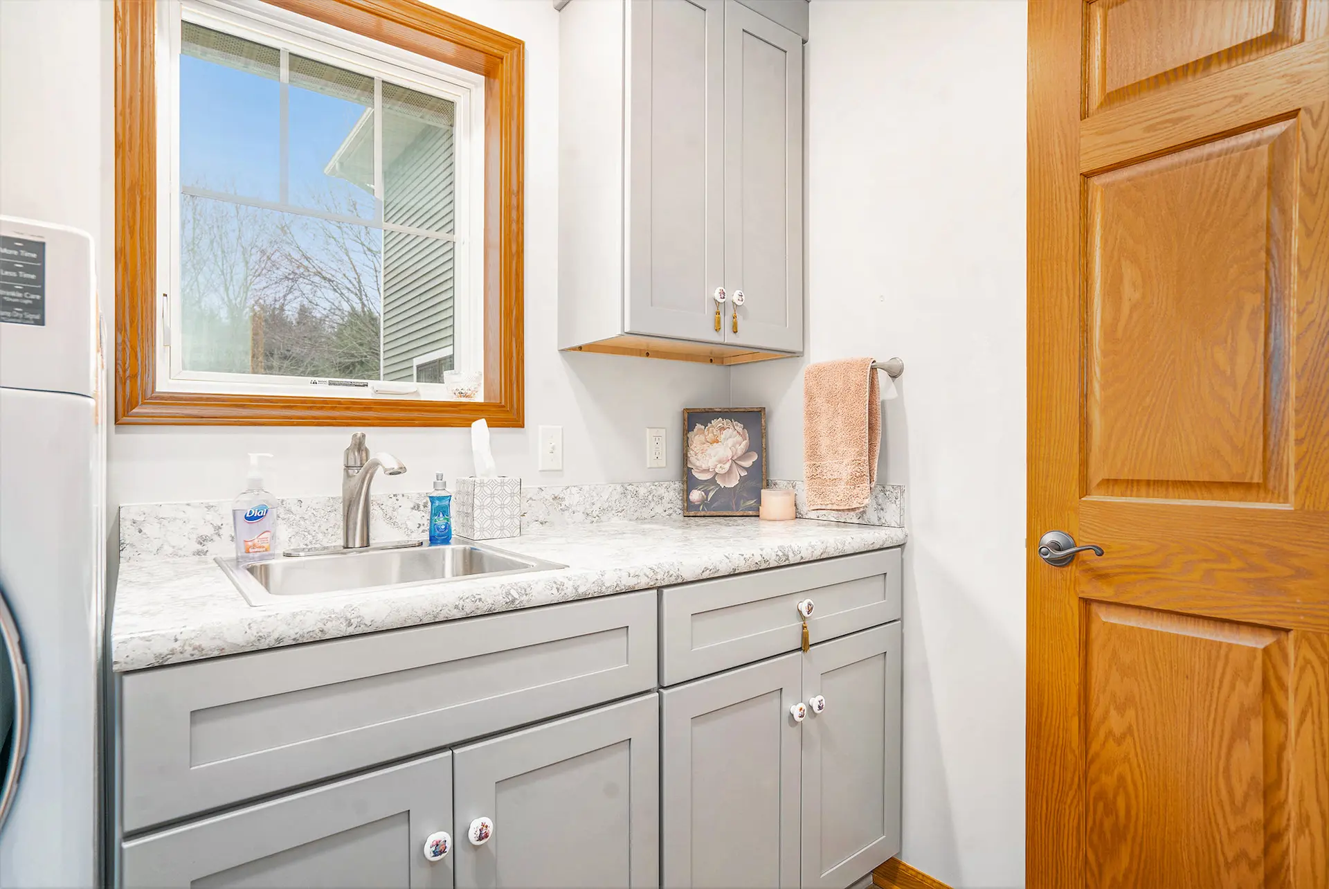 Laundry room utility sink with gray shaker cabinets, stone-look countertop, wood window trim, and wood door
