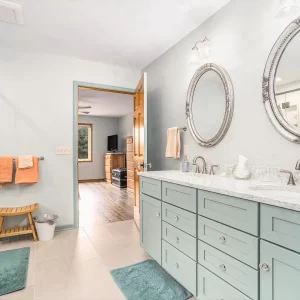 Bathroom with long double vanity, pale green cabinets, ornate round mirrors, glass shower enclosure, and light tile flooring