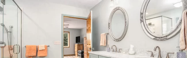Bathroom with Double Vanity and Glass Shower | Pennings & Sons Bathroom with long double vanity, pale green cabinets, ornate round mirrors, glass shower enclosure, and light tile flooring