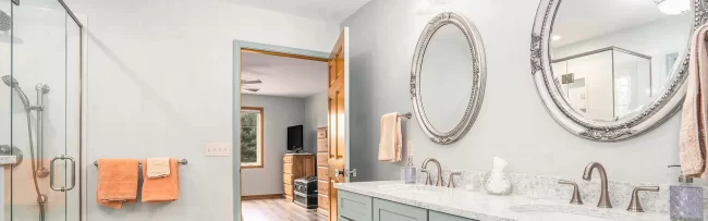 Bathroom with long double vanity, pale green cabinets, ornate round mirrors, glass shower enclosure, and light tile flooring