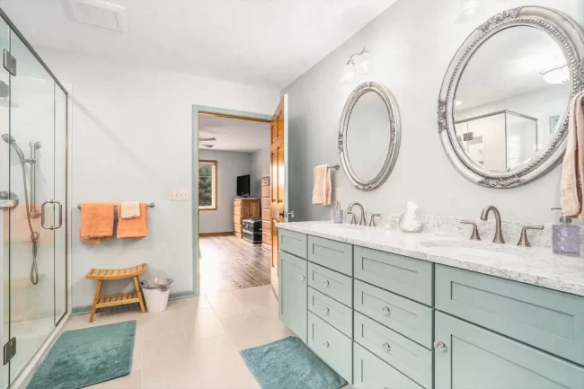 Bathroom with Double Vanity and Glass Shower | Pennings & Sons Bathroom with long double vanity, pale green cabinets, ornate round mirrors, glass shower enclosure, and light tile flooring