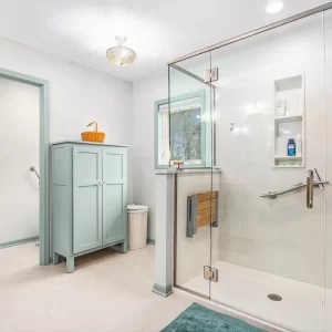 Spacious bathroom with glass shower enclosure, pale green storage cabinet, tile flooring, window, and soft blue-green trim