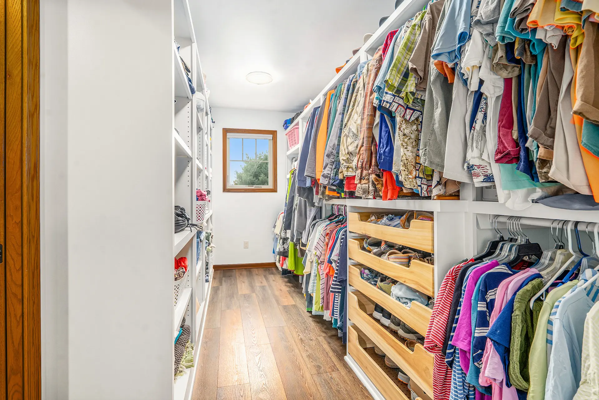 Walk-in closet with custom white shelving, hanging storage, wood flooring, shoe storage, and natural light from window