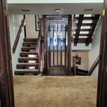 Custom residential elevator installed between two wood staircases with carpeted landing and glass lift enclosure