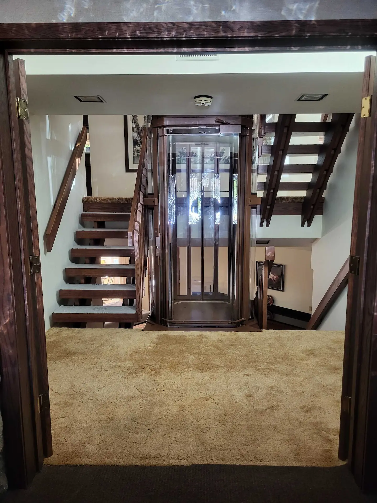 Custom residential elevator installed between two wood staircases with carpeted landing and glass lift enclosure