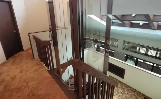 Glass Elevator Shaft and Stair Landing | Pennings & Sons Glass elevator shaft installed beside a wood staircase with warm carpet, railings, and multi-level access.