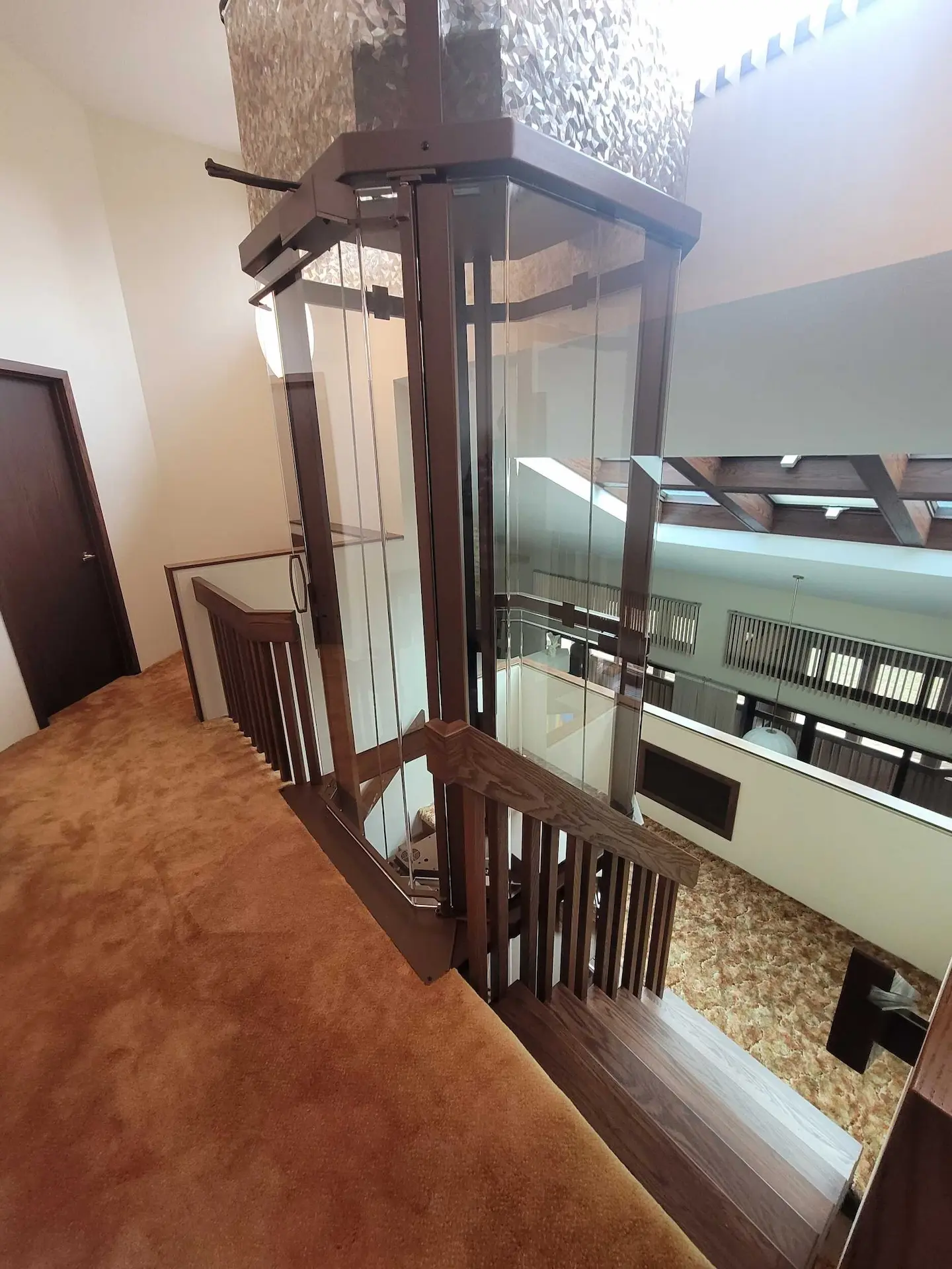 Glass elevator shaft installed beside a wood staircase with warm carpet, railings, and multi-level access.