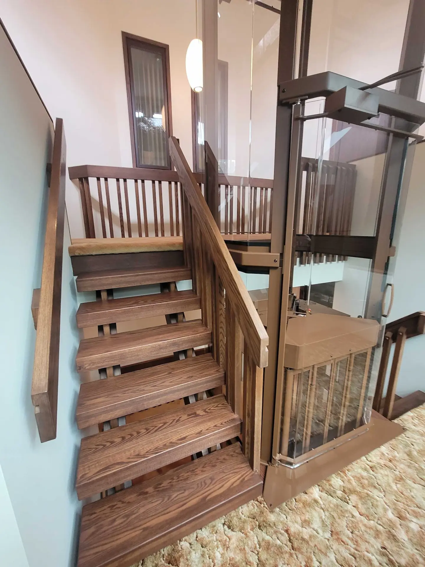 Home elevator positioned at a staircase landing with wood railings, glass panels, and carpeted floor area.