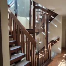 Custom residential elevator with clear glass panels positioned beside open wood stairs and matching railings