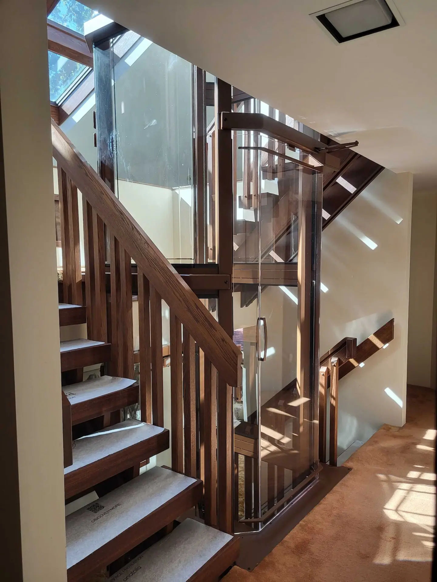 Custom residential elevator with clear glass panels positioned beside open wood stairs and matching railings