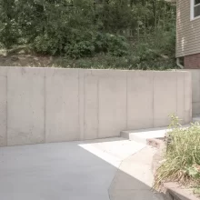 Finished concrete retaining wall and driveway area beside a rebuilt garage with landscaping and nearby home exterior