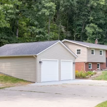 Completed garage rebuild connected to a split-level home with beige siding, white garage doors, new concrete driveway, and wooded backdrop