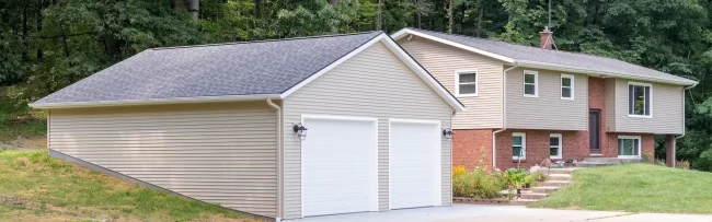 Completed garage rebuild connected to a split-level home with beige siding, white garage doors, new concrete driveway, and wooded backdrop
