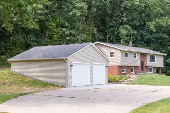Garage Rebuild with Home Exterior | Pennings & Sons Completed garage rebuild connected to a split-level home with beige siding, white garage doors, new concrete driveway, and wooded backdrop