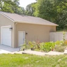 Side exterior view of a completed garage rebuild with beige siding, white garage doors, concrete drive, garden beds, and wooded hillside