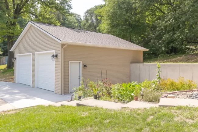 Garage Rebuild Side Exterior and Garden | Pennings & Sons Side exterior view of a completed garage rebuild with beige siding, white garage doors, concrete drive, garden beds, and wooded hillside