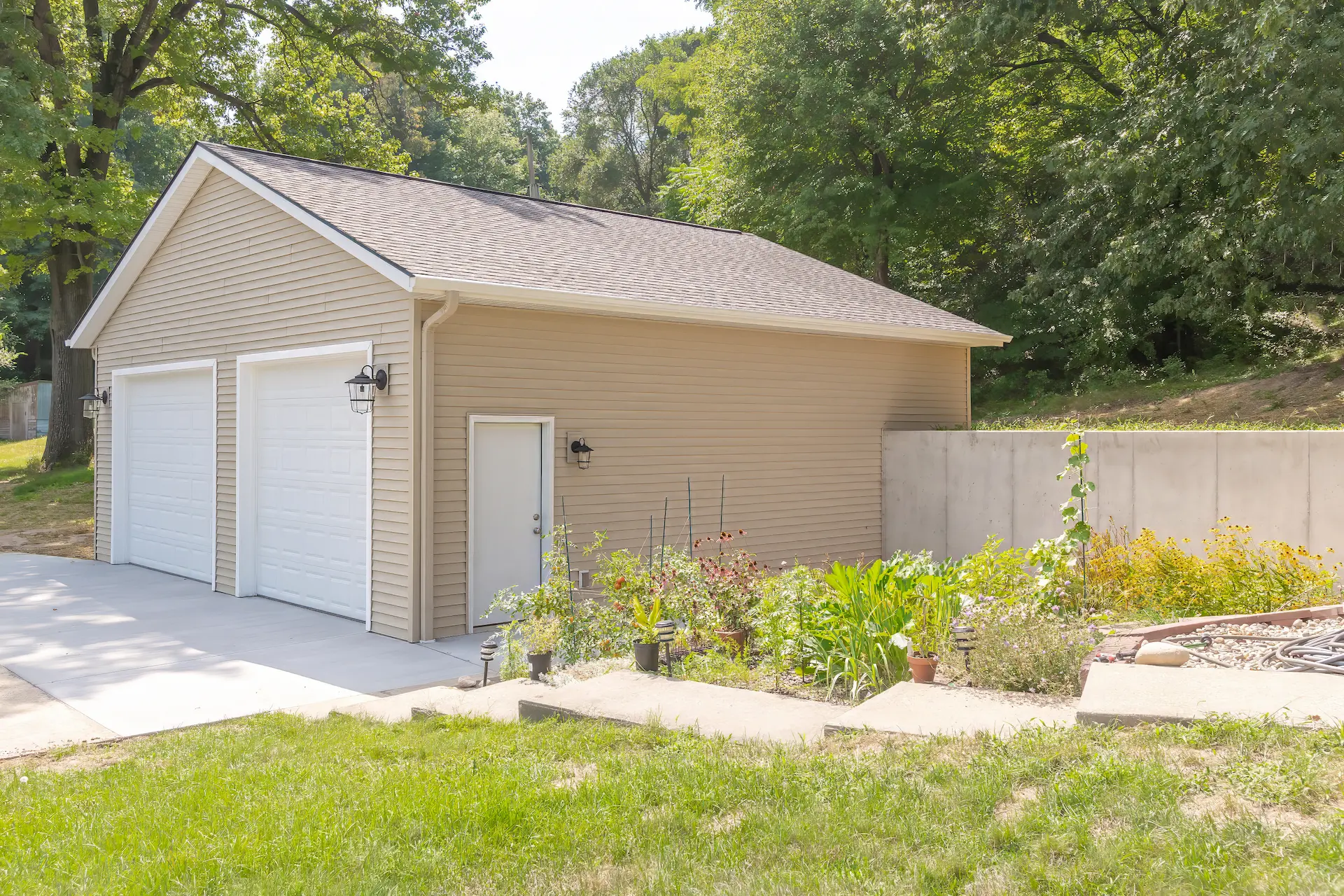 Side exterior view of a completed garage rebuild with beige siding, white garage doors, concrete drive, garden beds, and wooded hillside