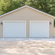 Front elevation of a completed two-car garage rebuild with beige siding, white overhead doors, exterior lights, and clean concrete driveway