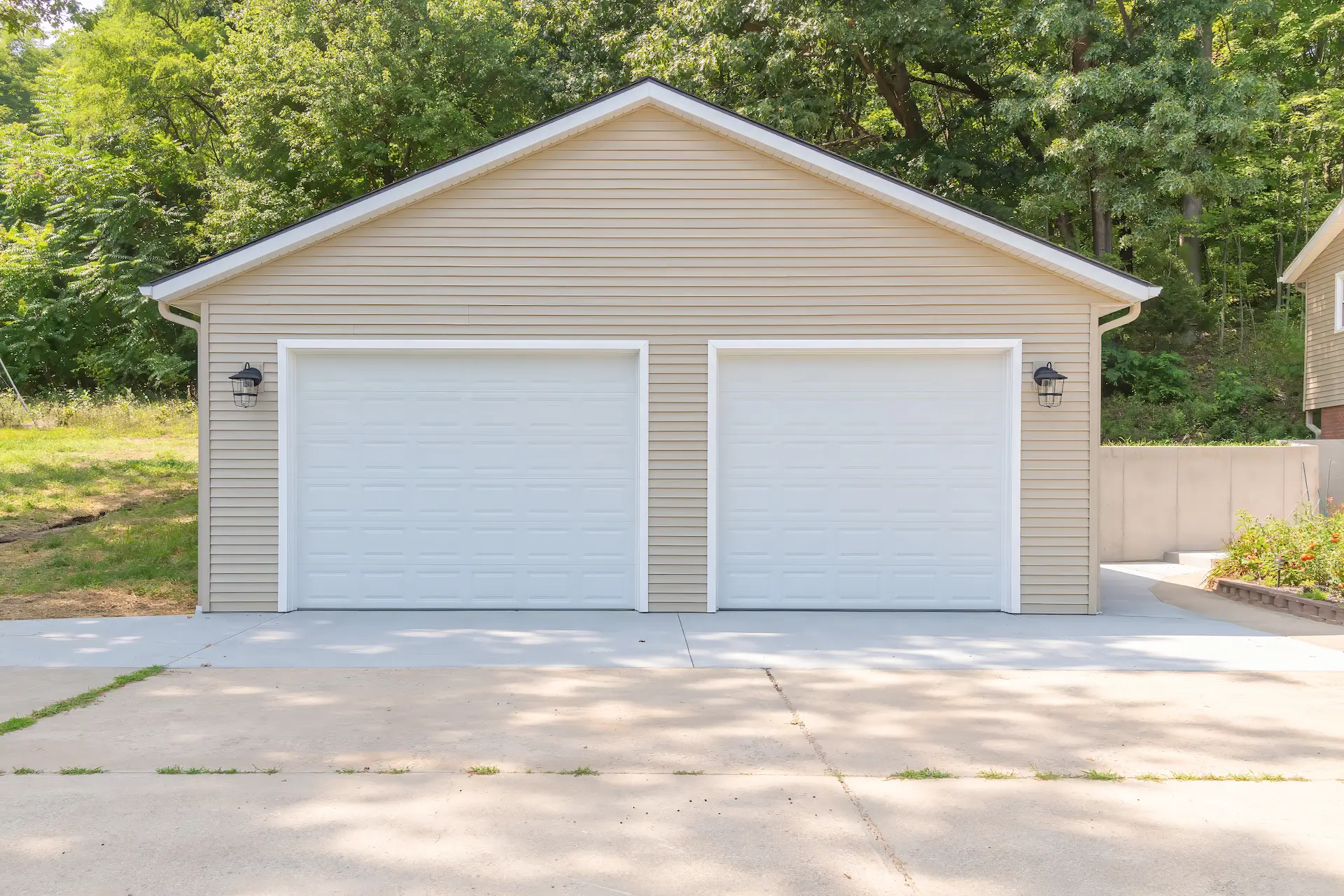 Front elevation of a completed two-car garage rebuild with beige siding, white overhead doors, exterior lights, and clean concrete driveway