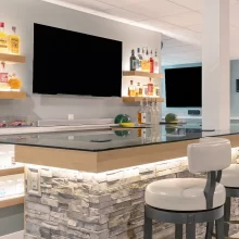 Basement bar area with floating bottle shelves, wall mounted TV, glass countertop, and display lighting