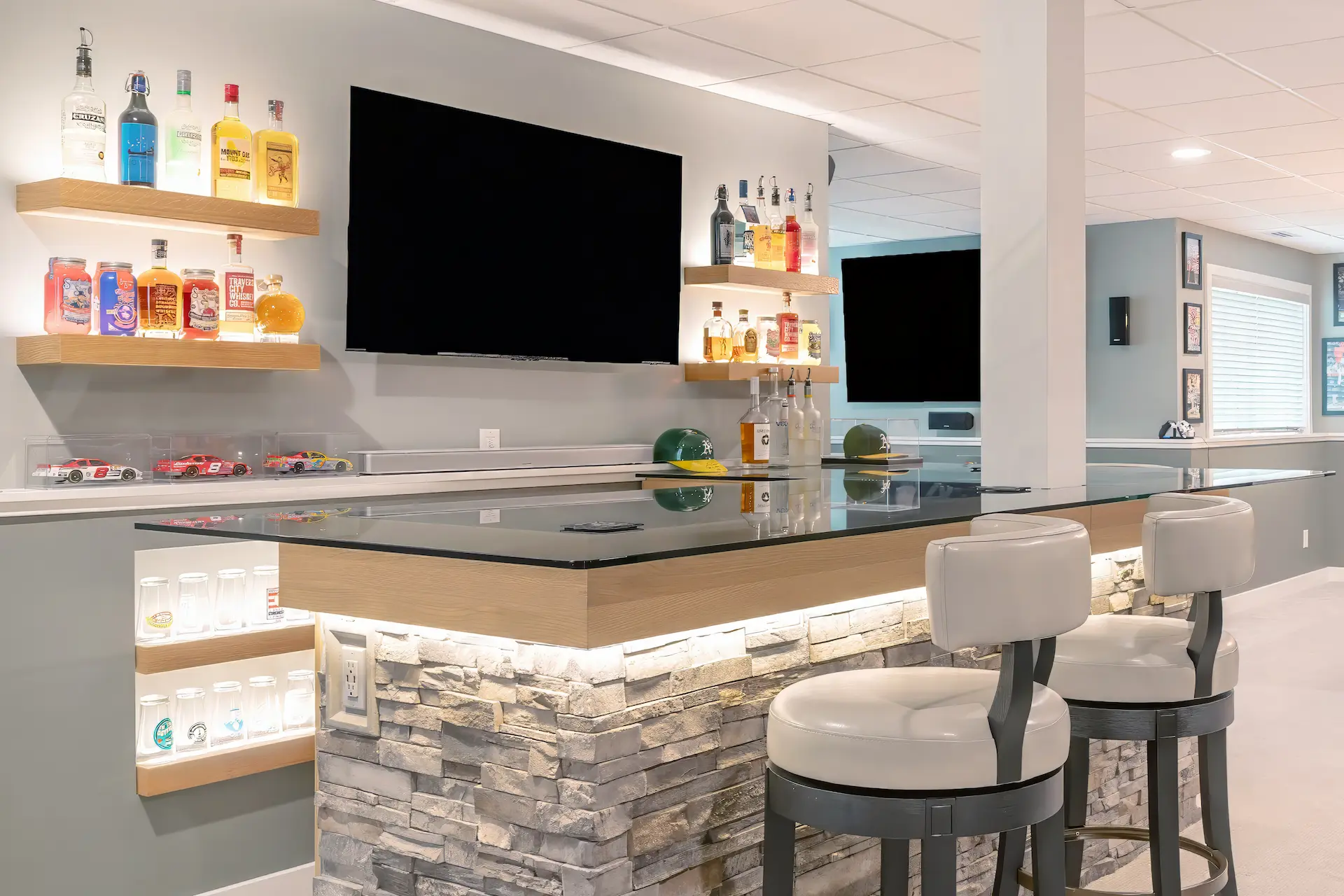 Basement bar area with floating bottle shelves, wall mounted TV, glass countertop, and display lighting