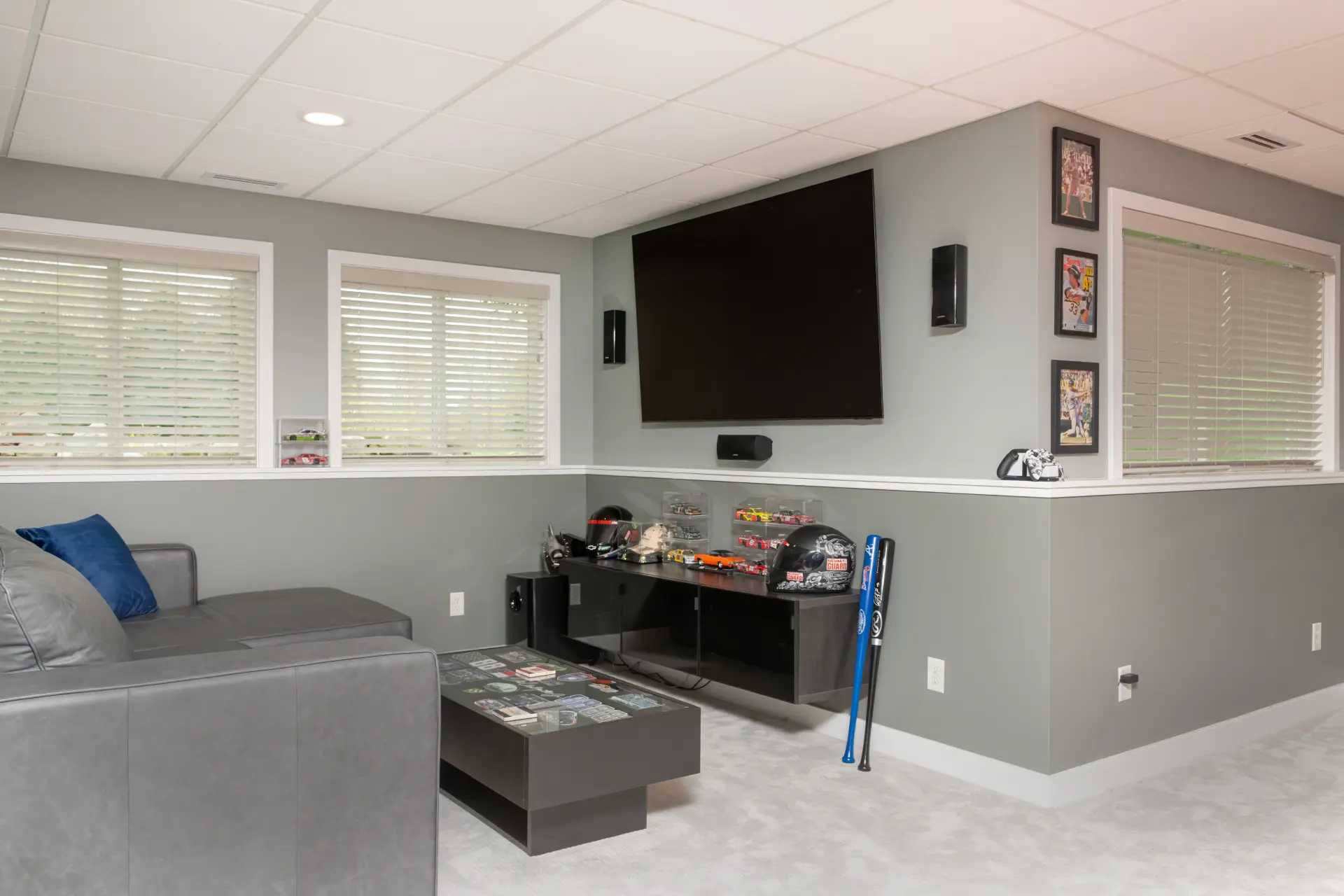 Basement media lounge with sectional seating, wall mounted TV, display shelves, and sports memorabilia