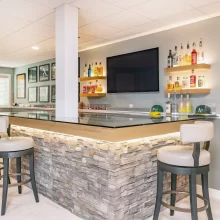 Custom basement bar with stone front, glass countertop, bar stools, floating shelves, and TV