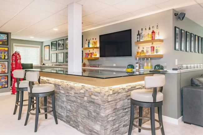 Custom Basement Bar with Stone Front | Pennings & Sons Custom basement bar with stone front, glass countertop, bar stools, floating shelves, and TV